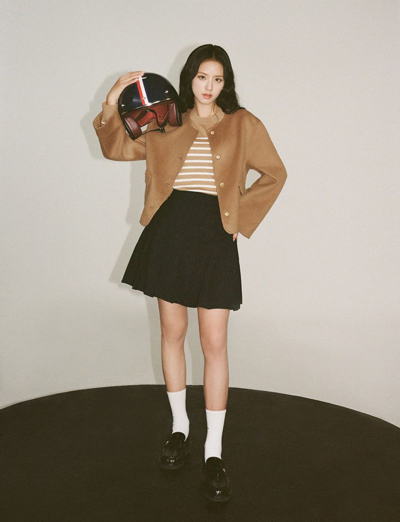 A fashionable woman poses in a studio, wearing a brown jacket and black skirt, holding a vintage motorcycle helmet with red and blue stripes.