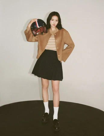A fashionable woman poses in a studio, wearing a brown jacket and black skirt, holding a vintage motorcycle helmet with red and blue stripes.