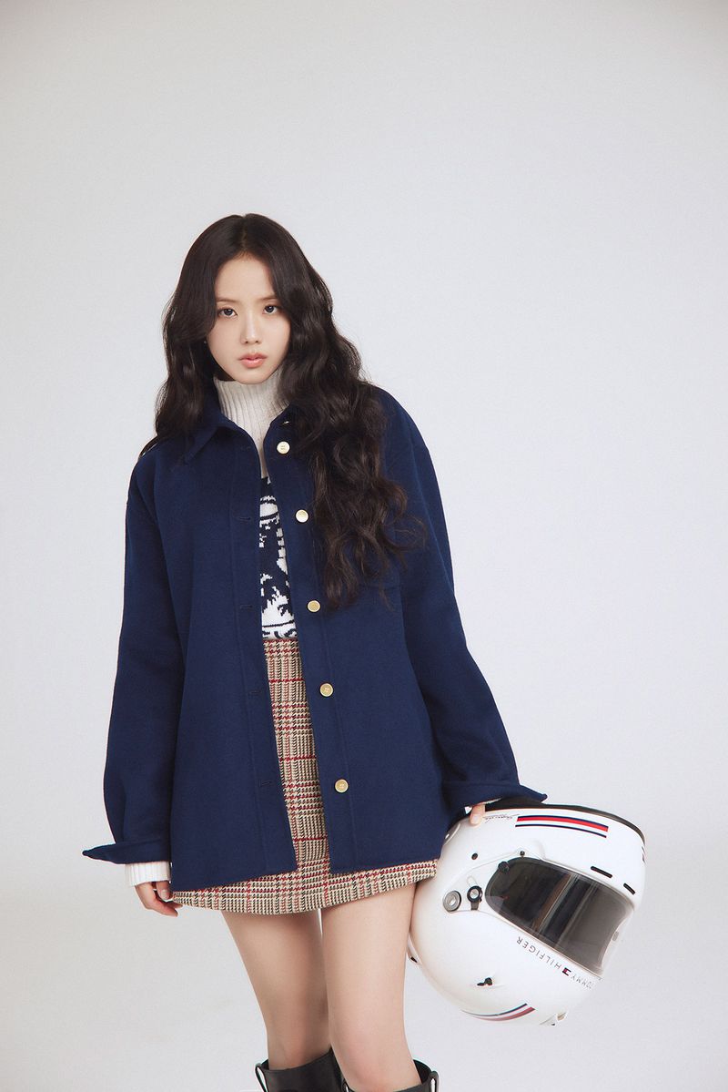 Young woman in blue jacket poses in a studio setting, holding a white racing helmet. She wears a skirt, and knee-high boots.