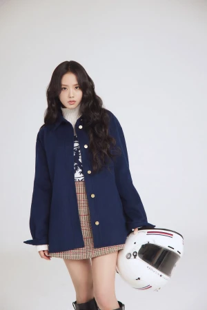 Young woman in blue jacket poses in a studio setting, holding a white racing helmet. She wears a skirt, and knee-high boots.