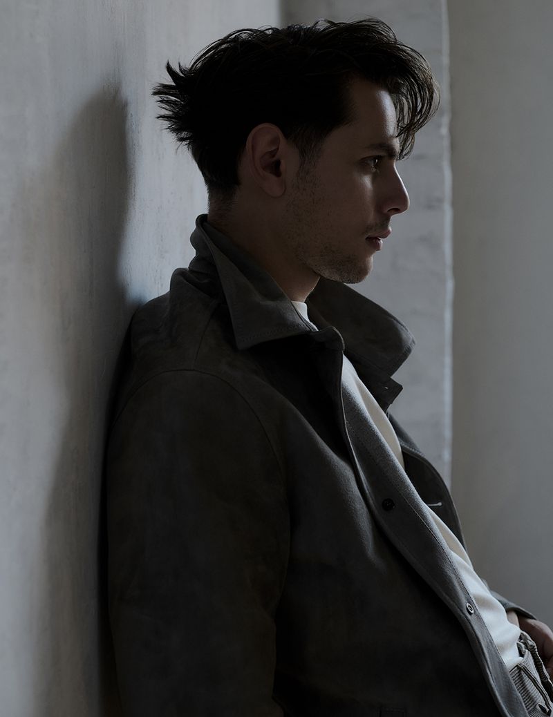 Portrait of a young adult man with dark hair in profile, wearing a stylish grey jacket, leaning against a neutral-toned wall in studio light.