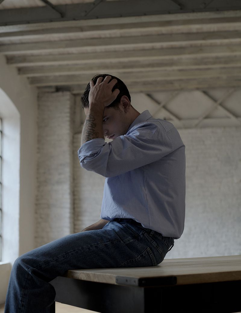 Adult man sits on a wooden table inside a room with his hands on his head wearing a blue button-down shirt and blue jeans.