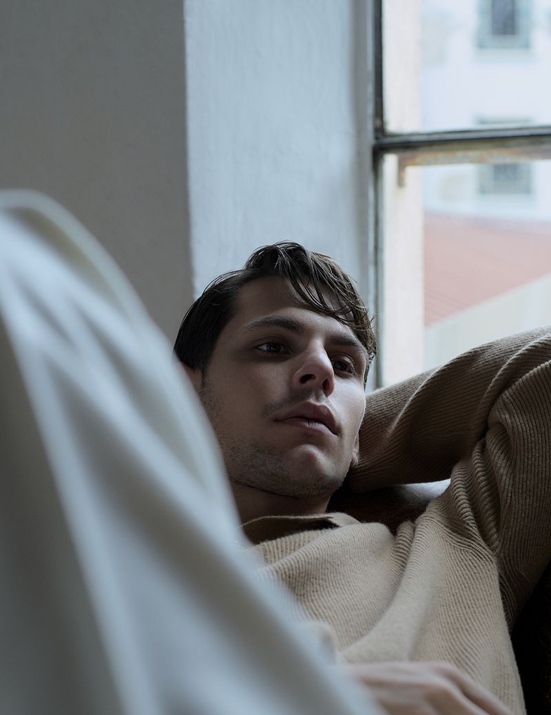 Portrait of a young adult man with dark hair in a beige sweater, reclining near a window with natural light, looking contemplative.