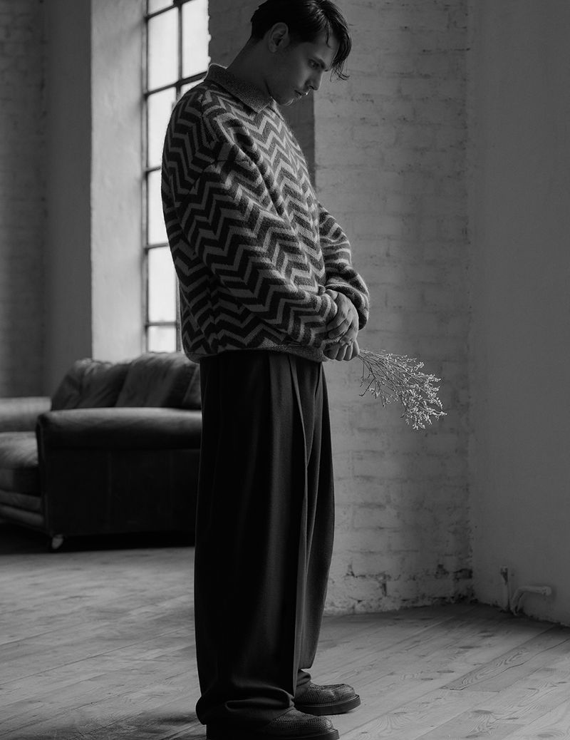 Monochrome portrait of a young adult male standing by a window, holding delicate foliage, with soft lighting and a textured brick wall.