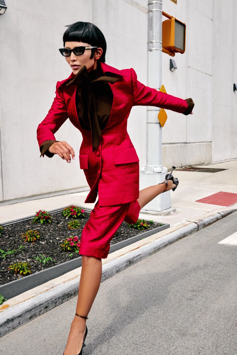 Woman in red suit runs across a crosswalk in an urban setting. The image features bright daylight and a dynamic pose.