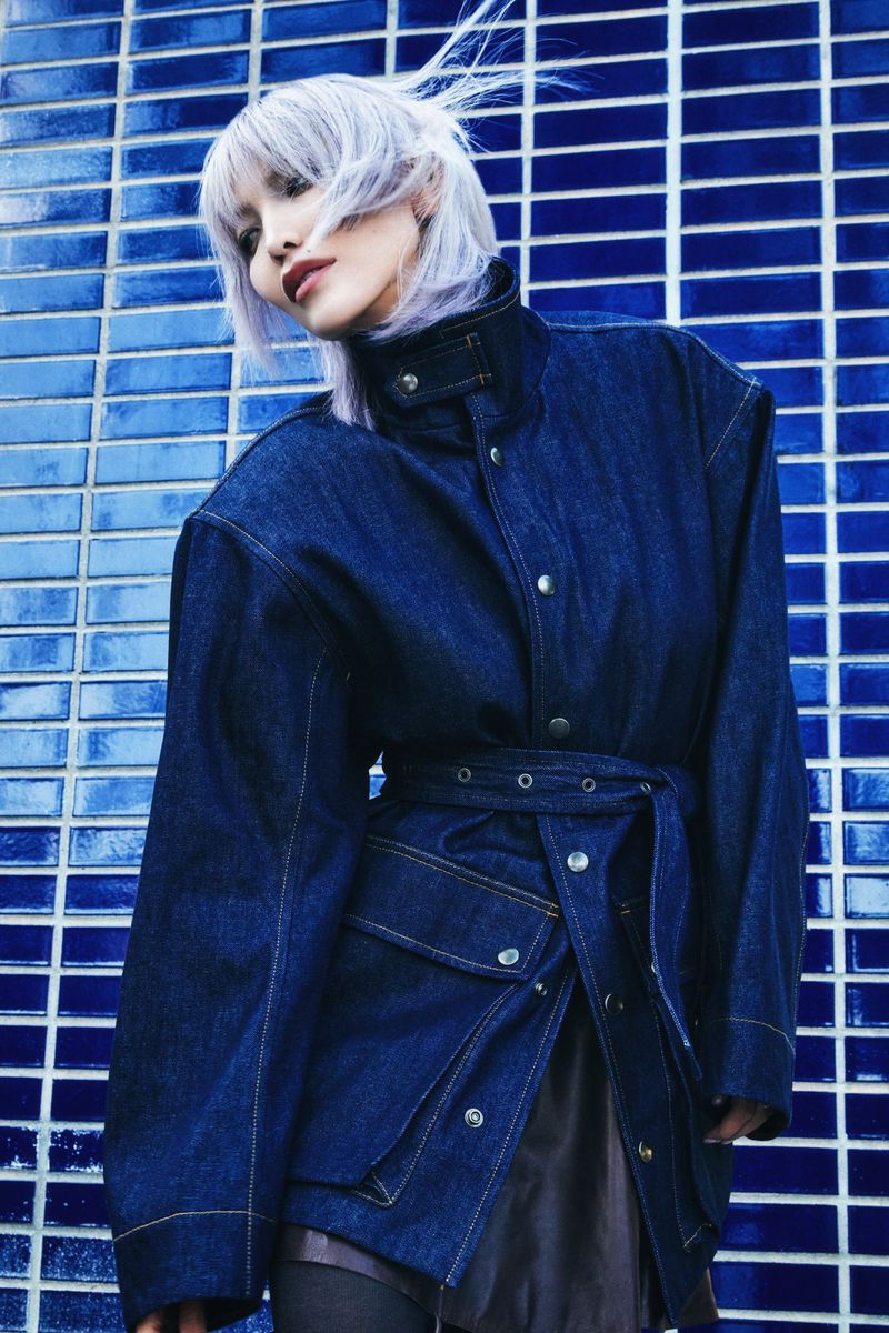 A stylish woman with silver hair poses in a belted denim jacket against a blue-tiled wall, showcasing a blend of fashion and urban backdrop.