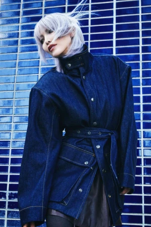 A stylish woman with silver hair poses in a belted denim jacket against a blue-tiled wall, showcasing a blend of fashion and urban backdrop.