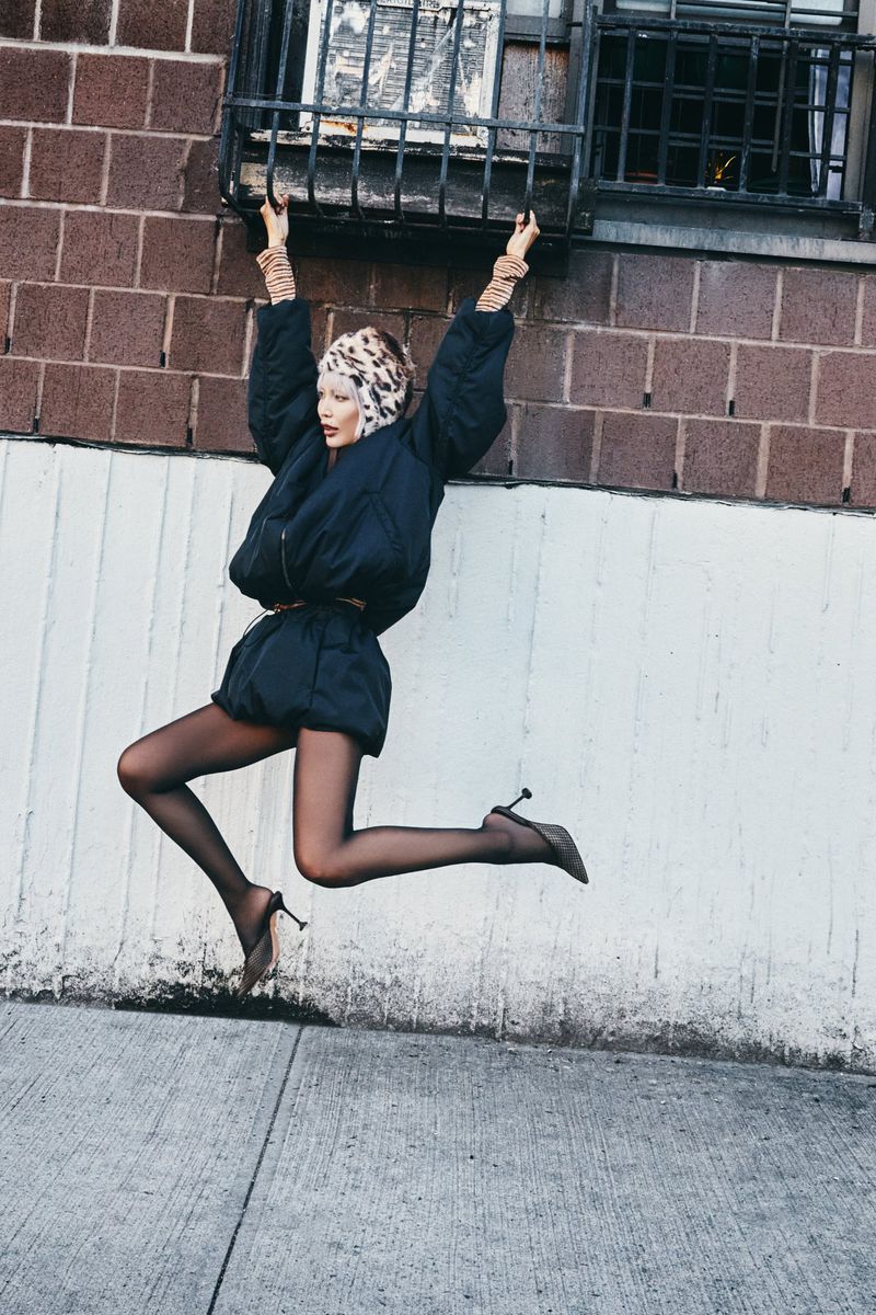 A slender adult woman in a black outfit, cheetah-print head covering, and stiletto heels jumps up toward window bars on a brick wall.