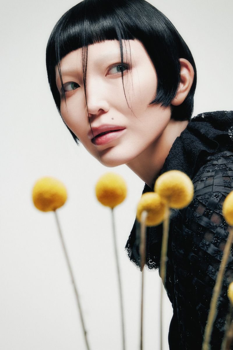 Woman with short black hair and yellow flowers. Portrait of a fair-skinned person with blunt bangs looking to the left.