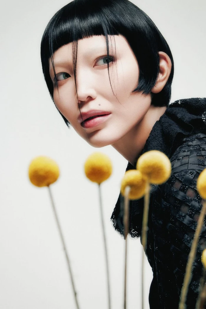 Close-up portrait of a woman with a dark bob haircut posing behind yellow billy button flowers against a white background.