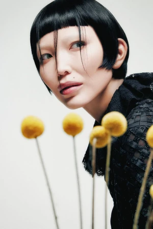 Close-up portrait of a woman with a dark bob haircut posing behind yellow billy button flowers against a white background.