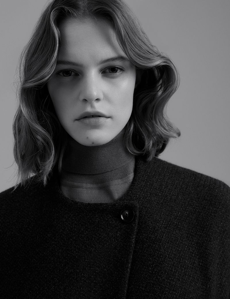 Black and white portrait of a woman with wavy hair wearing a turtleneck and dark textured wool coat against a plain backdrop.