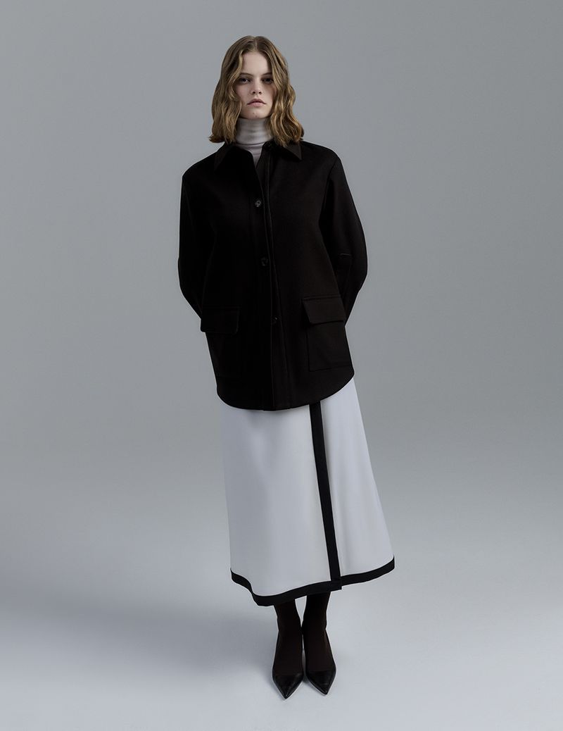 Fashionable woman in black jacket and white skirt stands in a photo studio, posing with hands behind her back and looking at the camera.