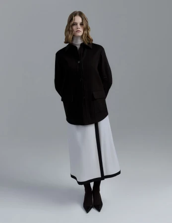 Fashionable woman in black jacket and white skirt stands in a photo studio, posing with hands behind her back and looking at the camera.