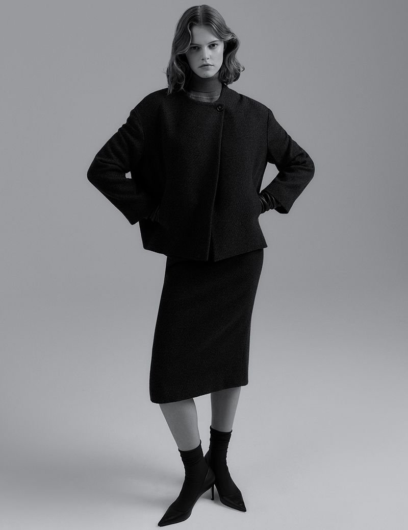 Adult female fashion model poses in a black short jacket and skirt in a studio with diffused lighting and a grey backdrop.
