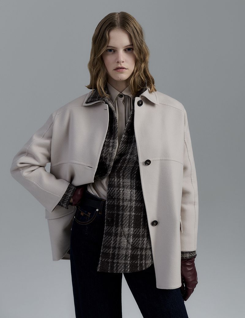 Woman in tan overcoat with plaid top, dark jeans, and leather gloves poses in front of a light gray backdrop in a studio setting.