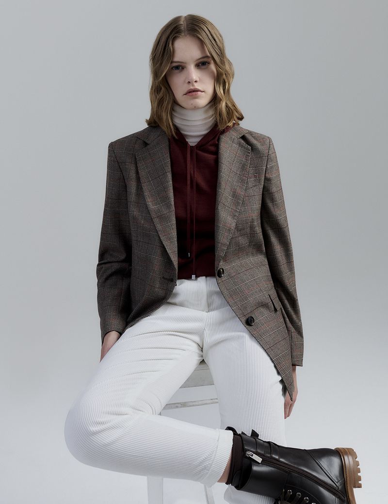 Young woman poses in a brown plaid blazer, white pants, and black boots against a neutral background in a fashion-forward studio shot.