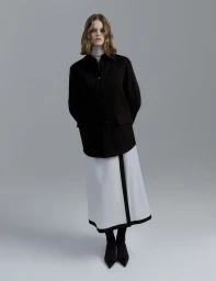 Woman wearing an oversized black jacket and a white skirt with black trim standing against a grey studio backdrop.