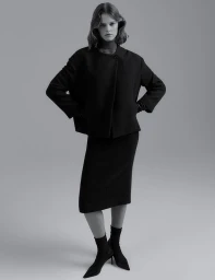 Model in dark jacket and midi skirt with hands on hips against a plain grey background