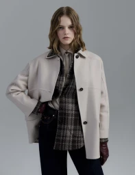 Model in an off-white wool coat, plaid shirt, and dark trousers against a grey seamless background