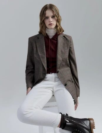 Fashion model seated in a brown plaid blazer, white ribbed trousers, and dark combat boots against a neutral grey studio background.