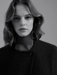 Black and white portrait of a woman with wavy hair wearing a turtleneck and dark textured wool coat against a plain backdrop.