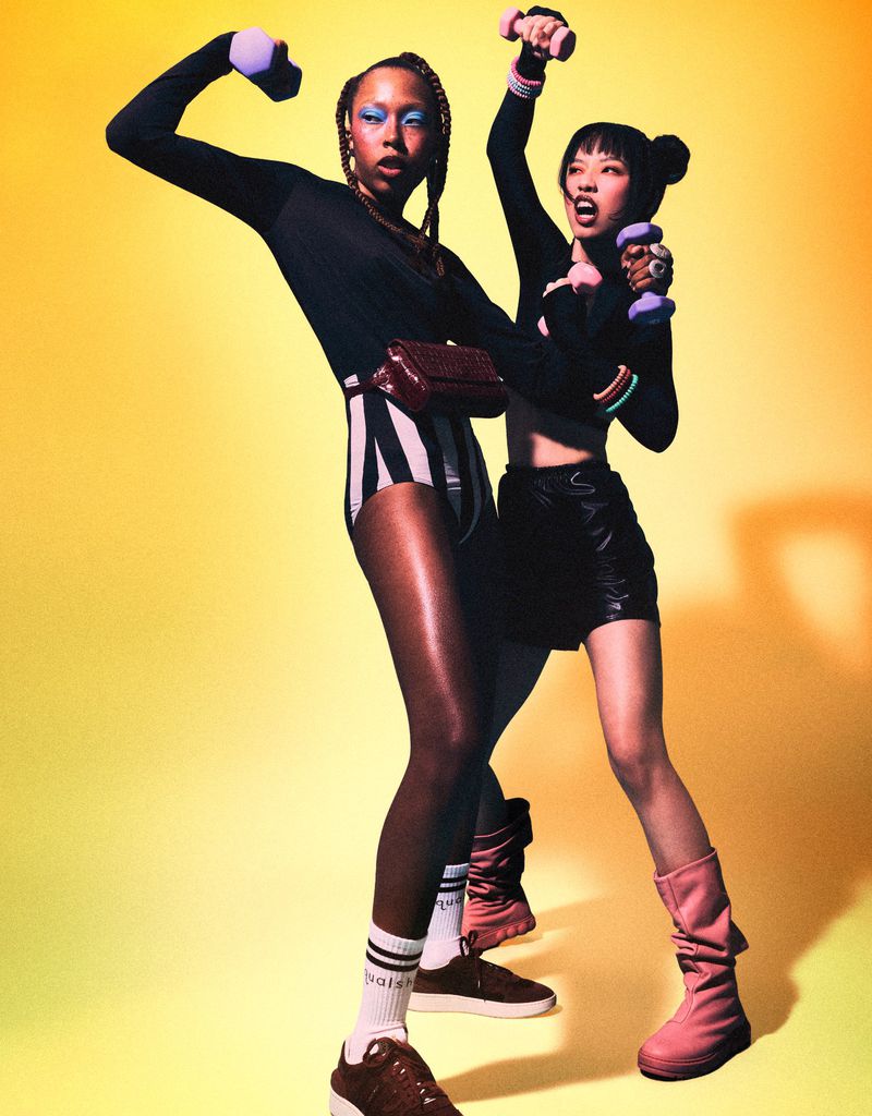 Two athletic women in sportswear hold dumbbells in a vibrant studio. One wears striped shorts and the other a black skirt.