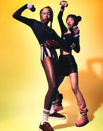 Two athletic women in sportswear hold dumbbells in a vibrant studio. One wears striped shorts and the other a black skirt.