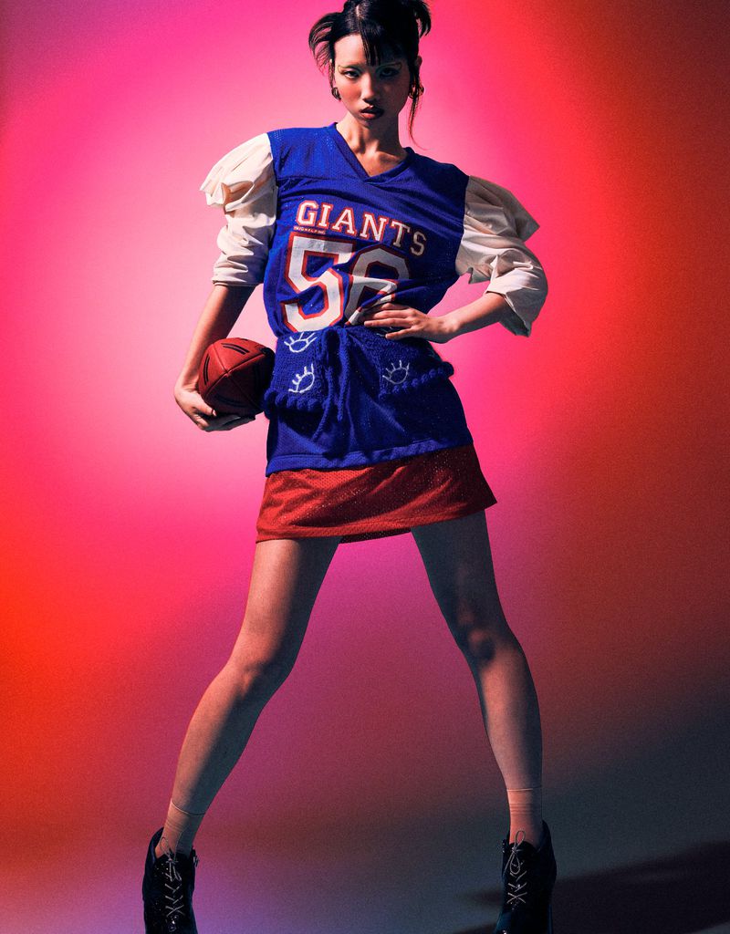 A young woman poses in an athletic-inspired outfit, holding a football against a vibrant red and pink gradient background in a fashion forward image.