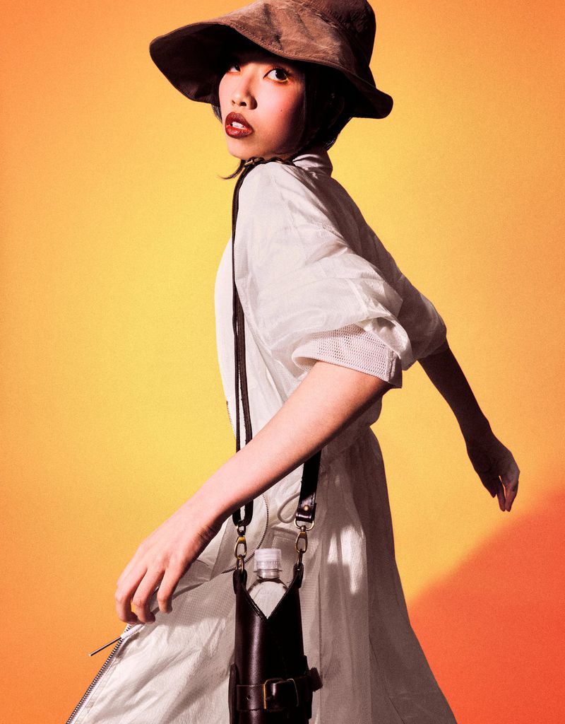 A young woman in a hat and white dress carries a water bottle in a leather holder against a blended yellow-orange background.