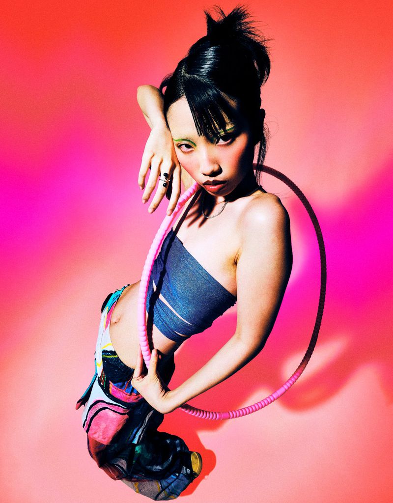 Young Asian woman poses with a hula hoop against a vibrant pink gradient background in a studio setting. Unique fashion and makeup.