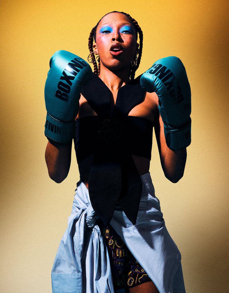 Adult female boxer in studio wearing turquoise boxing gloves, a black top, and a light blue skirt, striking a confident pose.