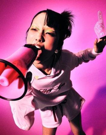 Asian woman shouting into a pink megaphone and holding a #1 foam finger against a vibrant pink background in a close-up shot.