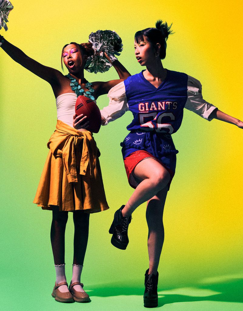 Two young women strike a pose in a studio with a yellow-green backdrop. One holds a football and pom-poms. Both wear sporty outfits.