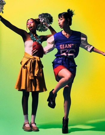 Two young women strike a pose in a studio with a yellow-green backdrop. One holds a football and pom-poms. Both wear sporty outfits.