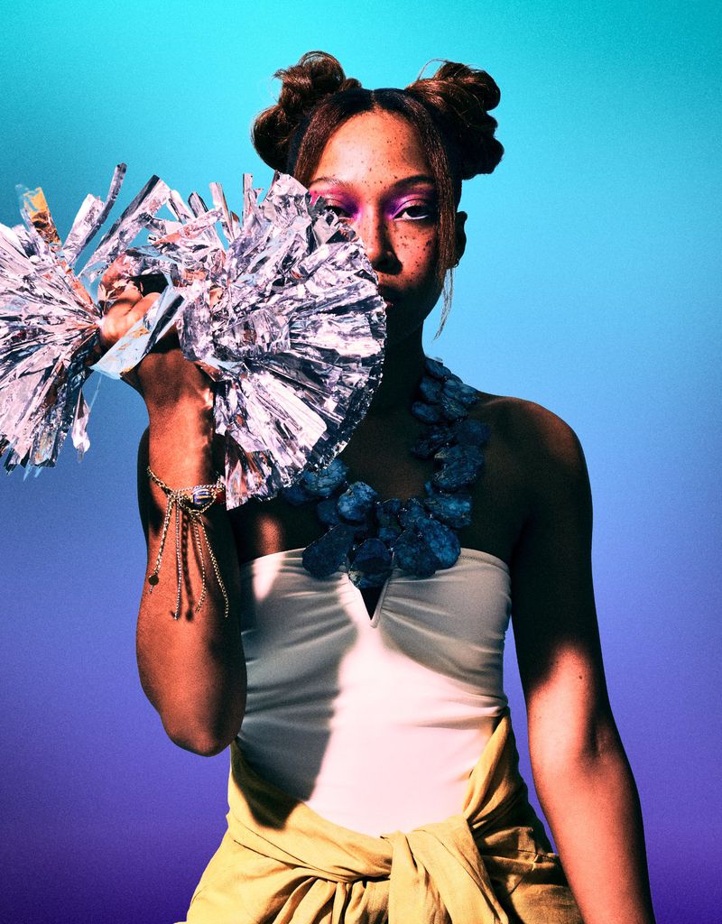 A young adult woman with two space buns and silver pom-poms against a gradient blue and purple background in a studio setting.