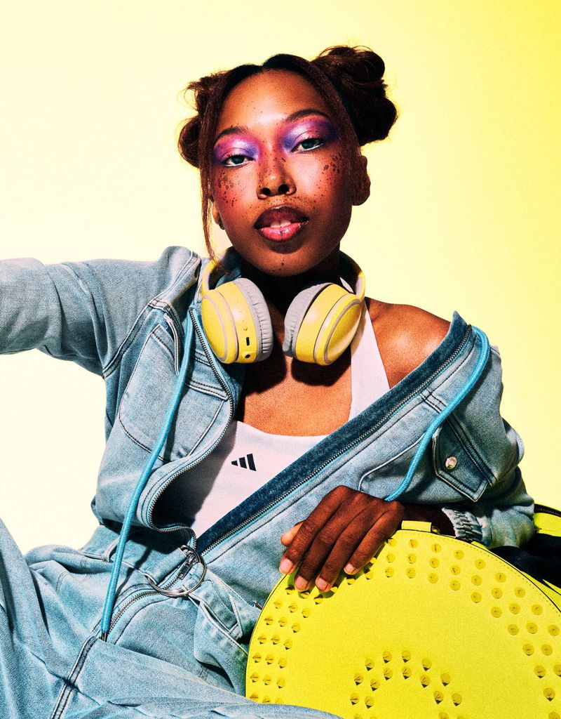 Stylish young woman with space buns, headphones, and a denim jacket poses against a yellow background, holding a perforated yellow disc.