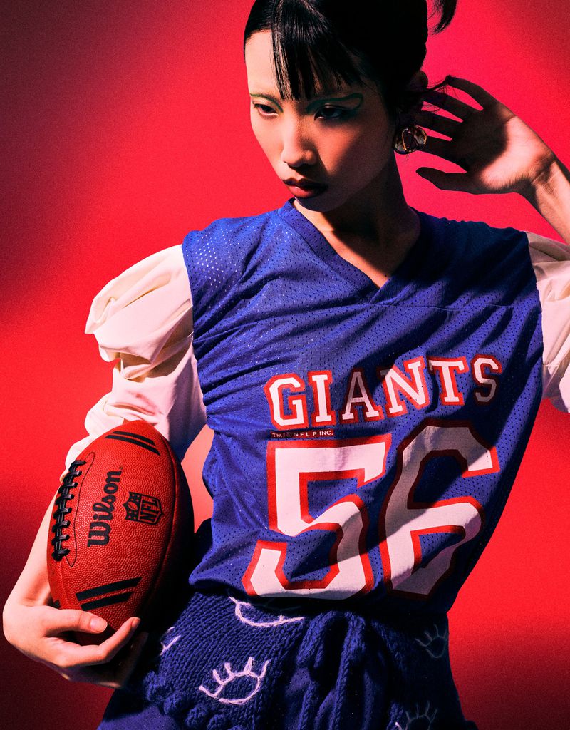 Portrait of an Asian woman in a Giants jersey holding a football, posing against a vibrant red backdrop in a studio setting.