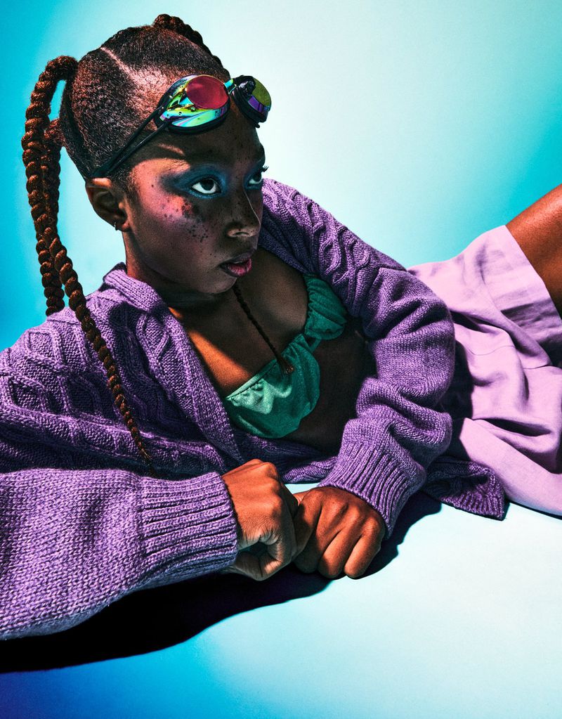Young person lying down, wearing goggles and a cardigan, illuminated by blue light, showcasing a studio portrait with creative style.