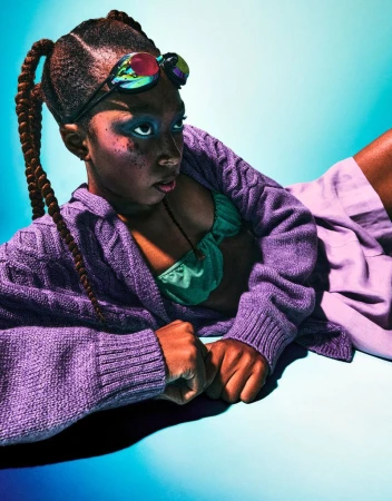 Young person lying down, wearing goggles and a cardigan, illuminated by blue light, showcasing a studio portrait with creative style.