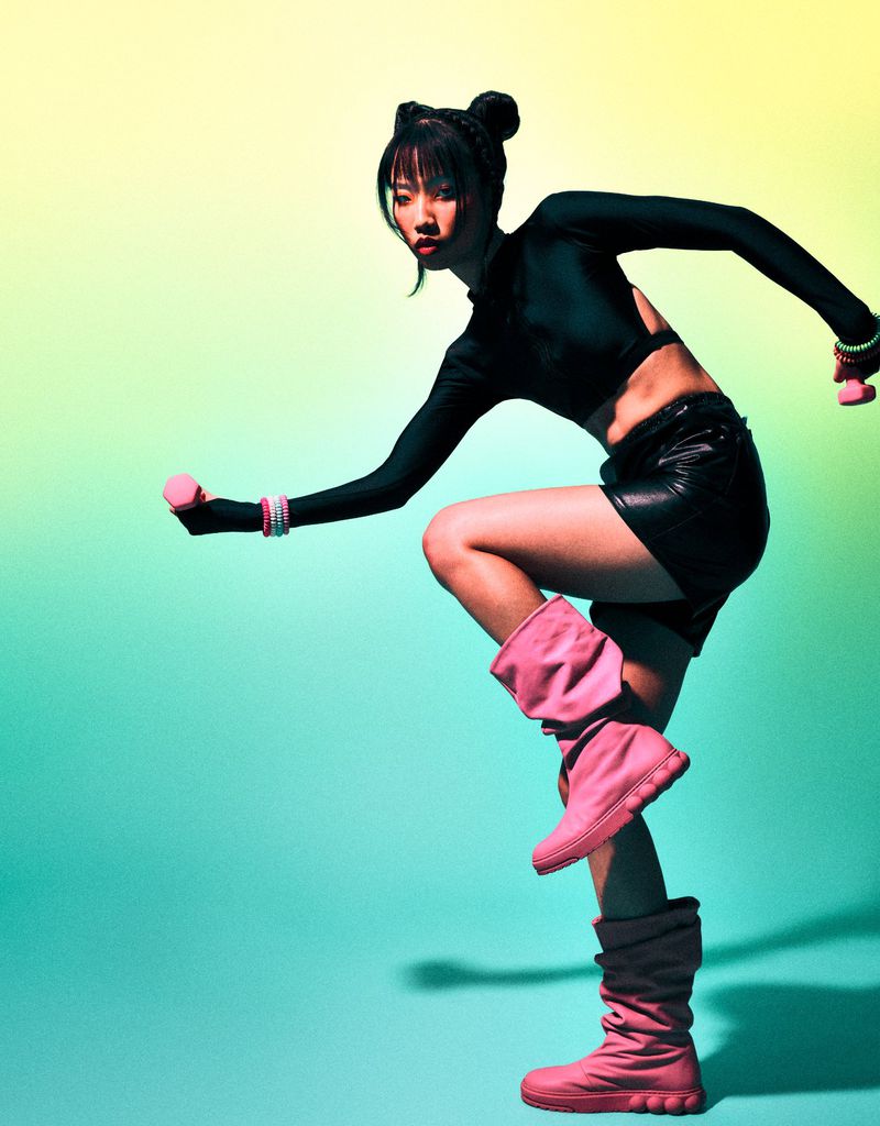 Athletic Asian woman in black sportswear and pink boots, exercising with pink dumbbells on a graduated yellow to blue background in a studio.