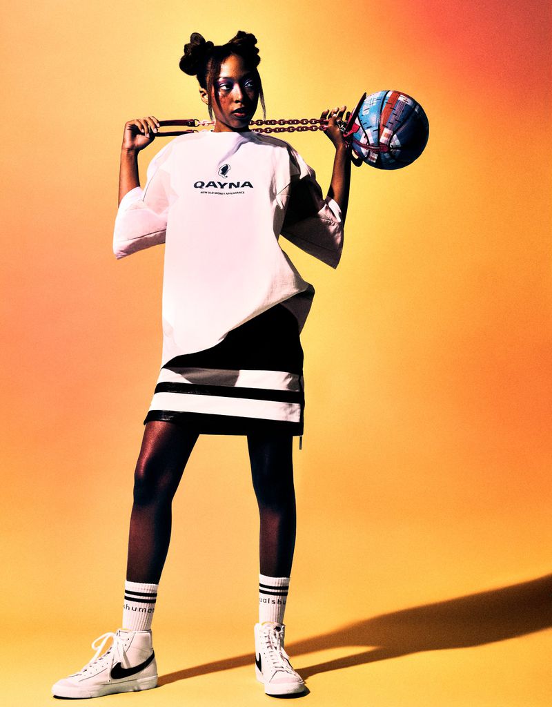 A woman in streetwear fashion poses in a studio with a basketball attached to a chain, in front of a graduated orange background.