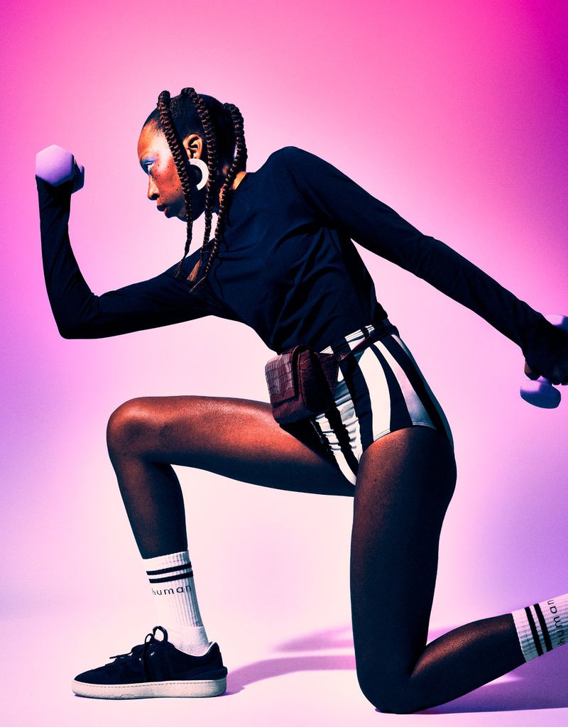 Athletic woman in striped shorts kneels in a studio against a pink background, holding dumbbells during a fitness workout session.