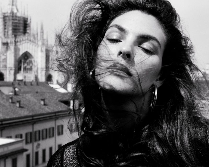 Black and white portrait of a woman with windblown hair and eyes closed, set against a backdrop of European architecture and city buildings.