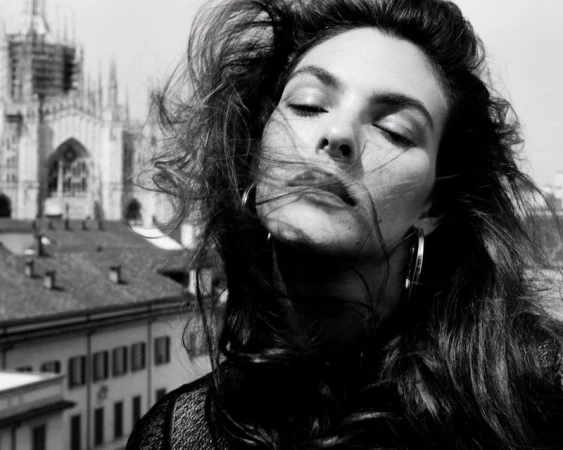 Black and white portrait of a woman with windblown hair and eyes closed, set against a backdrop of European architecture and city buildings.