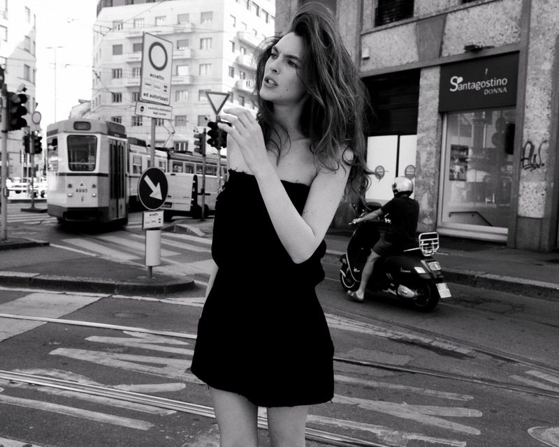 Monochrome portrait of a young woman in a dress standing on a city street with a tram and buildings in the background.