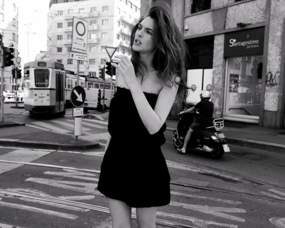 Monochrome portrait of a young woman in a dress standing on a city street with a tram and buildings in the background.