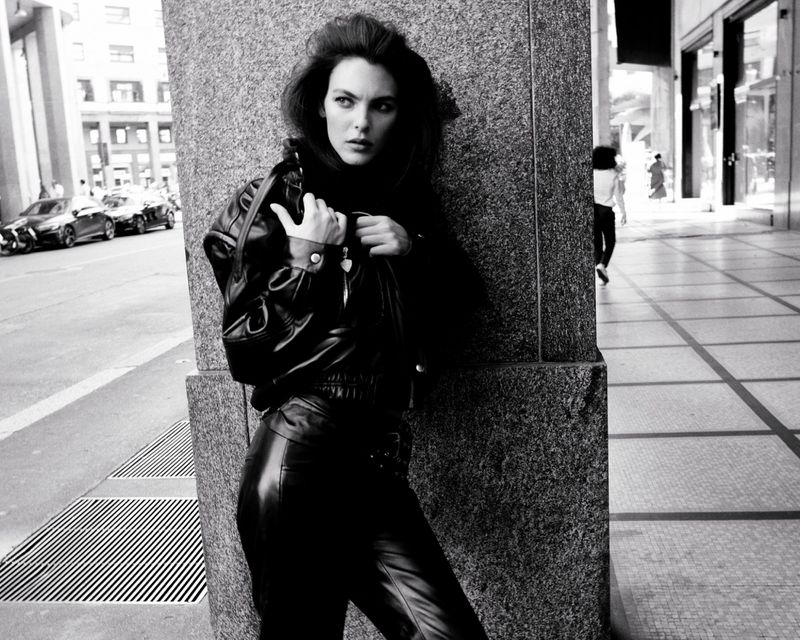 Model Vittoria Ceretti leaning against a stone pillar on a city street wearing a black leather jacket and trousers