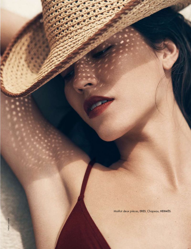 Close-up of a woman in a dark red bathing suit and a woven hat that casts shadows on her face, eyes closed, and red lipstick.