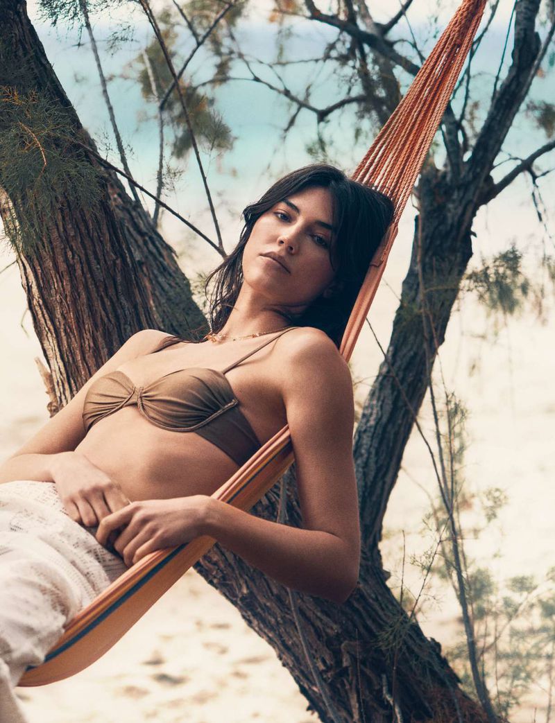 Adult woman relaxing in an orange hammock suspended between trees on a beach, wearing a brown bikini top and white linen pants.